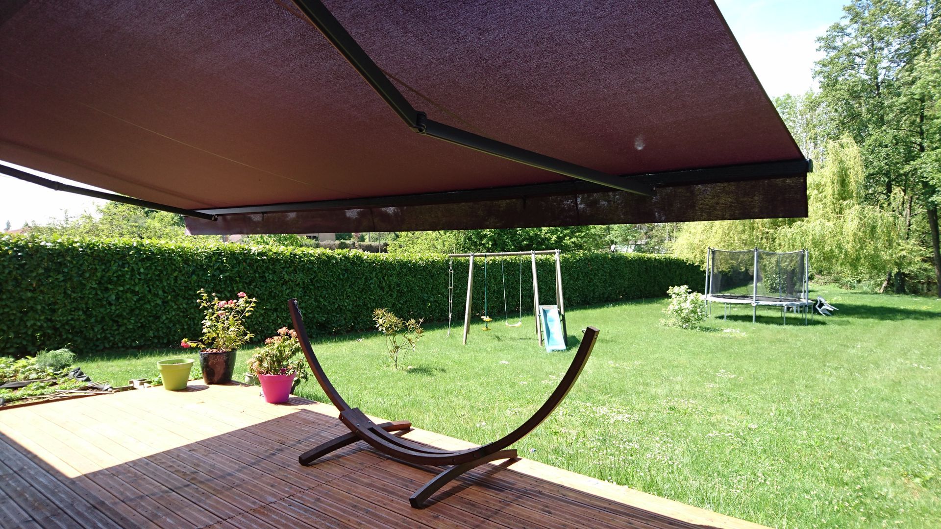 Scène extérieure avec un auvent marron au-dessus d'une terrasse en bois, un hamac et une pelouse verte arborée.