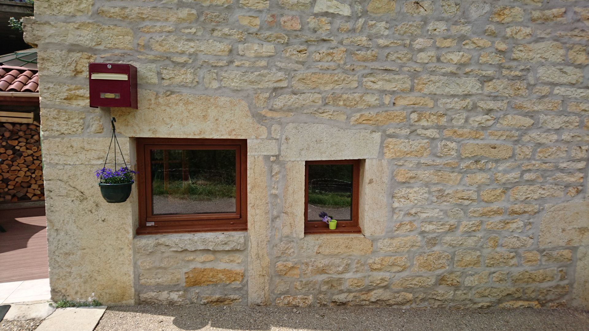Façade en pierre d'un bâtiment avec deux fenêtres, une boîte aux lettres et un pot de fleurs suspendu.