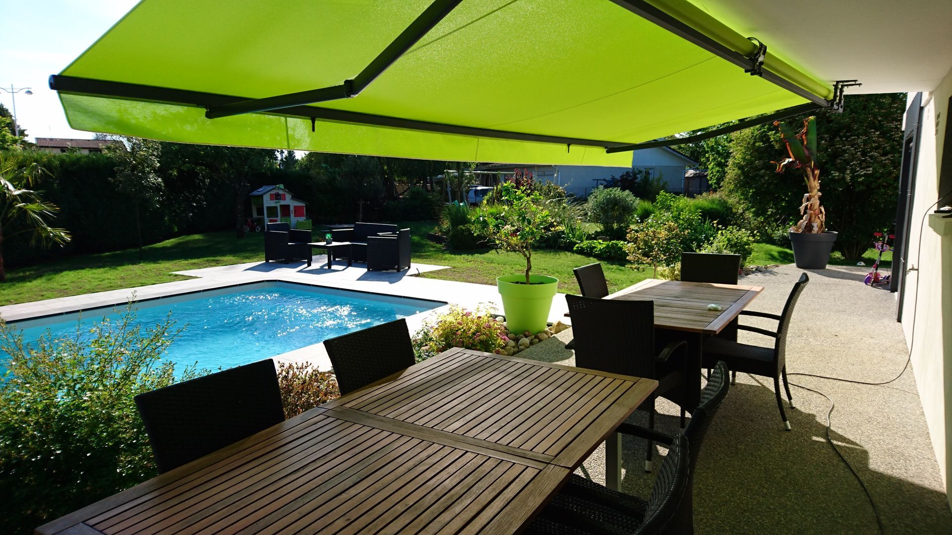 Une terrasse dans le jardin avec un auvent vert au-dessus d'une table à manger, une piscine et de la verdure.