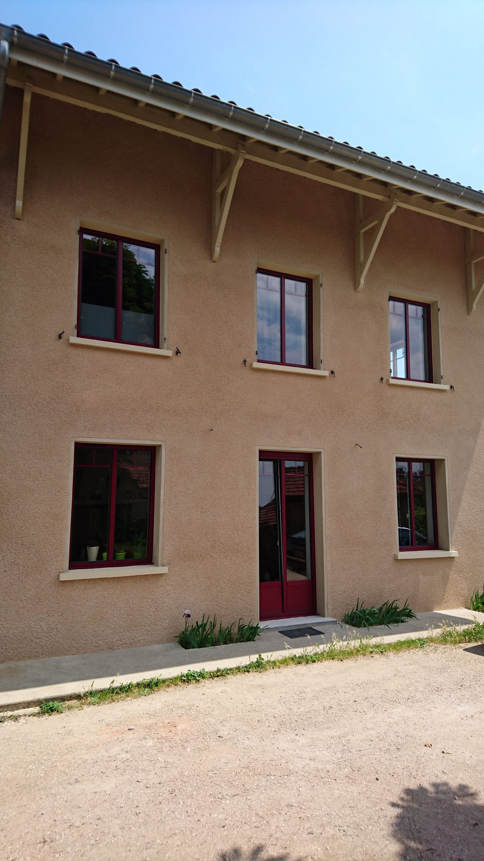 Bâtiment beige de deux étages avec fenêtres et porte à encadrement rouge, sous un ciel bleu.