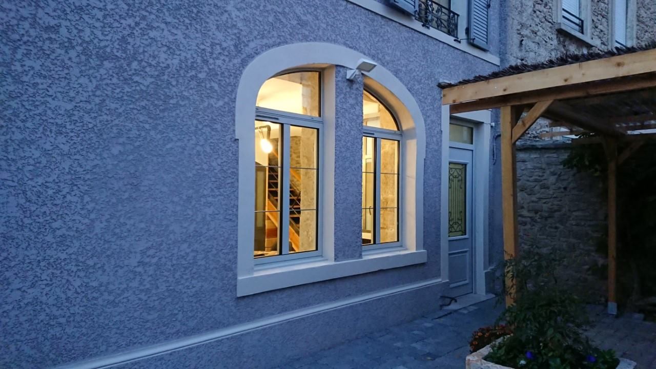 Bâtiment en stuc bleu avec des fenêtres cintrées, éclairées de l'intérieur, à côté d'une porte et d'une pergola en bois.