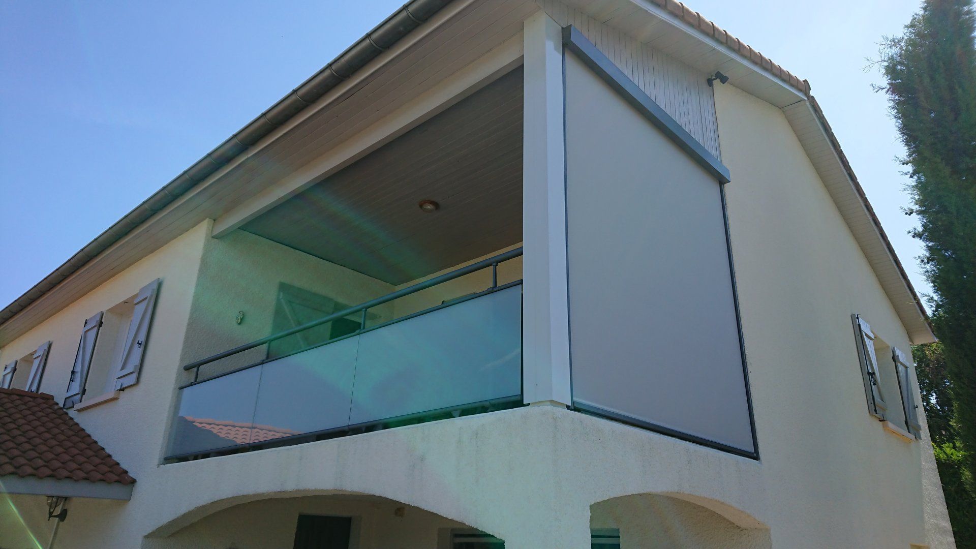 Maison beige avec un balcon aux tons gris, une rambarde en verre et un ciel bleu.