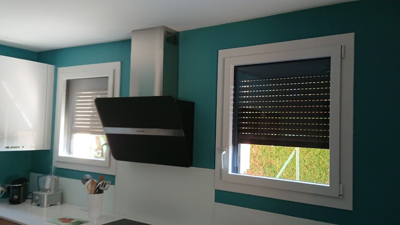 Cuisine aux murs turquoise, placards blancs et fenêtres équipées de stores enrouleurs foncés ; hotte aspirante centrée.