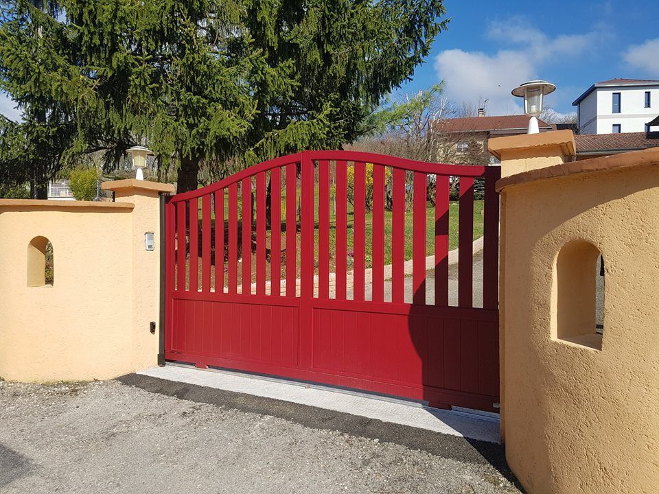 Portail rouge entre deux piliers beiges, allée en béton, maisons en arrière-plan.