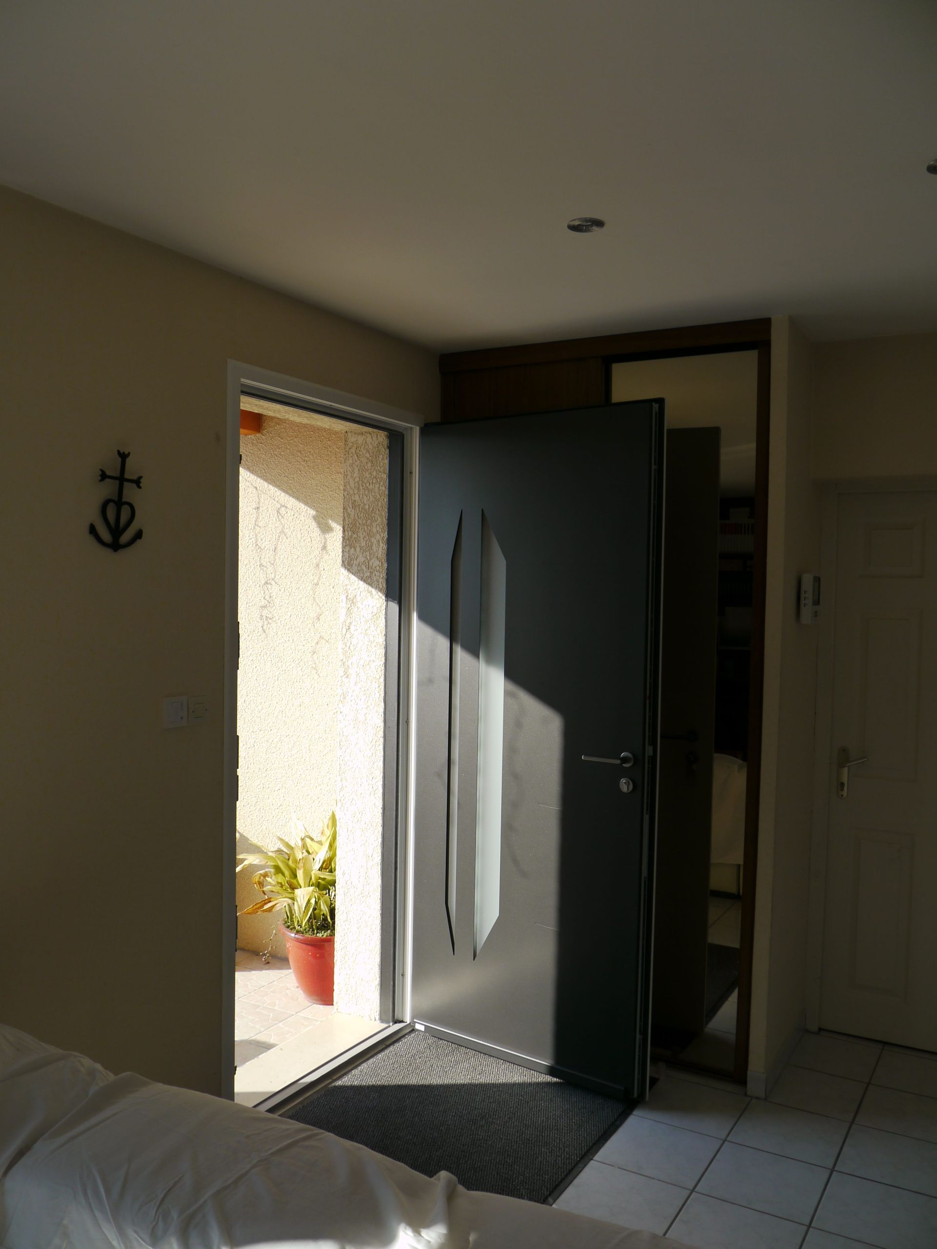 Porte grise ouverte, lumière du soleil à l'intérieur. Entrée décorée d'ancres au mur et d'une plante en pot.