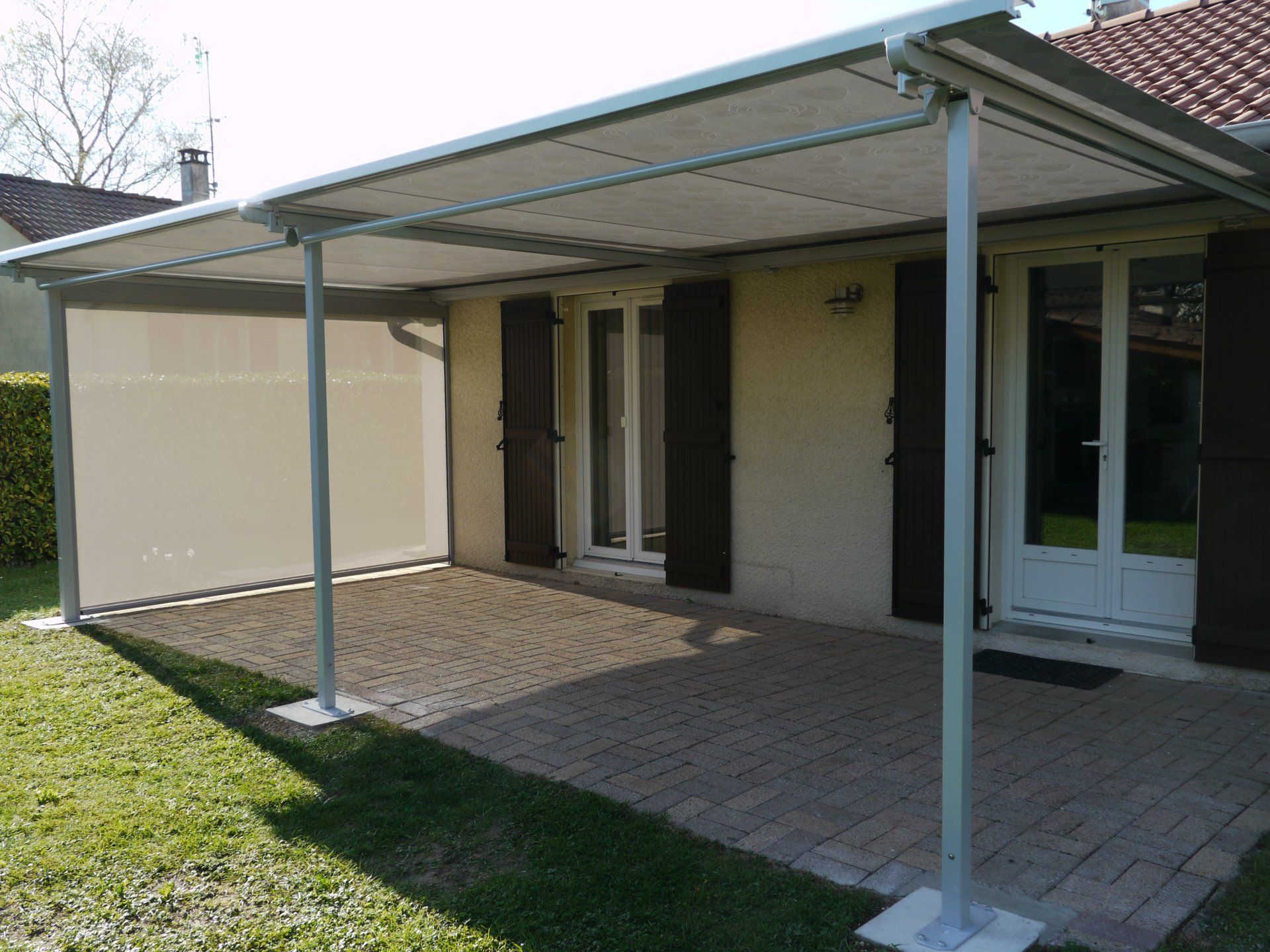 Auvent de terrasse à structure métallique fixé à une maison beige, avec terrasse en pierre et pelouse.