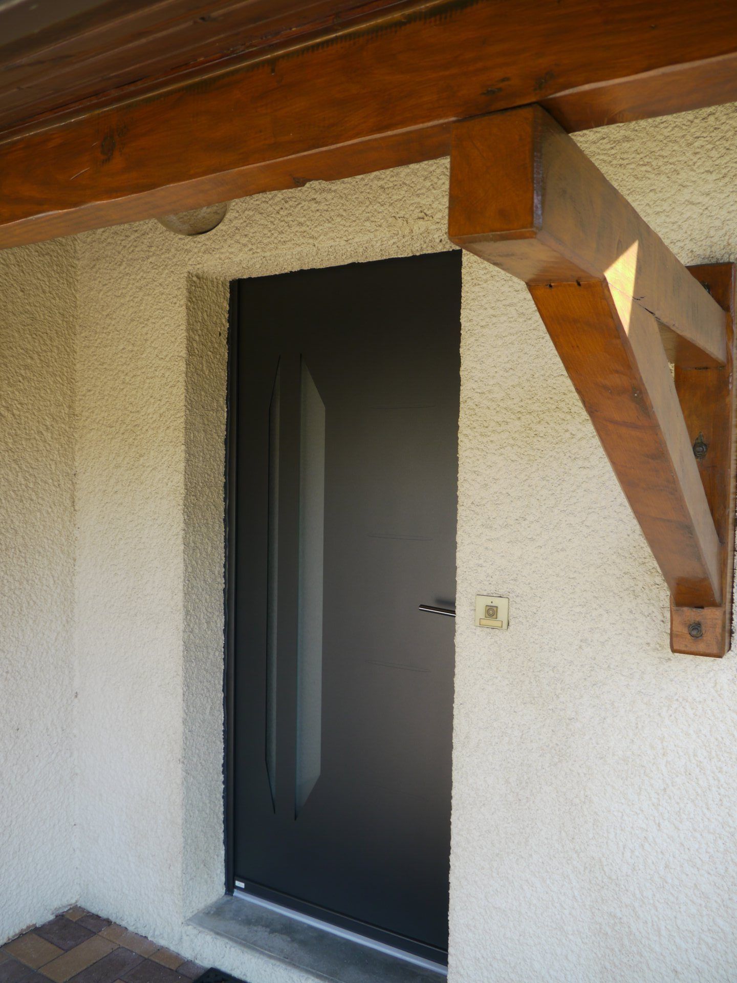Porte d'entrée noire sous une poutre en bois, mur en stuc beige.
