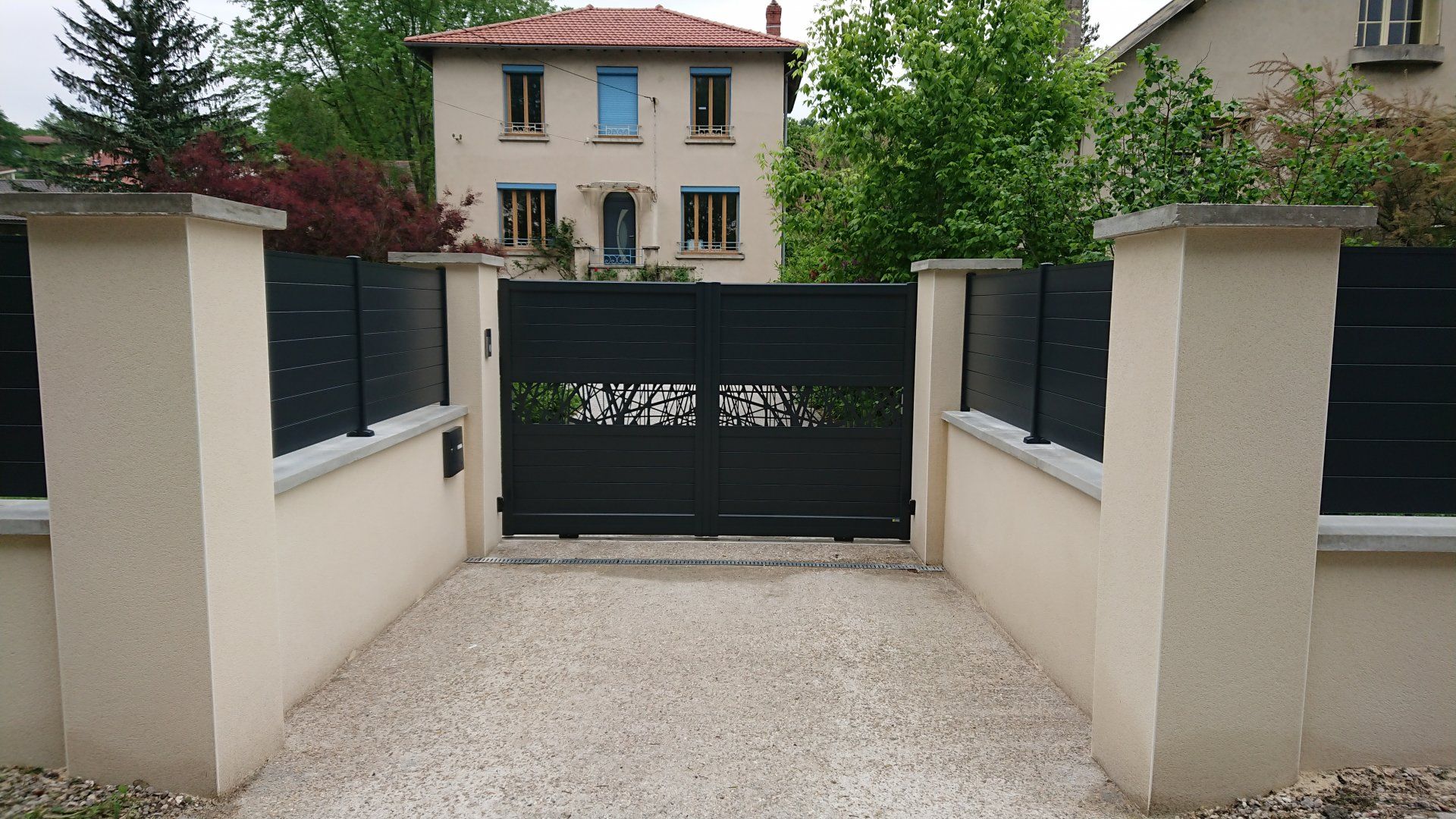 Portail noir à motifs décoratifs, entre piliers et murs beiges, allée de gravier.