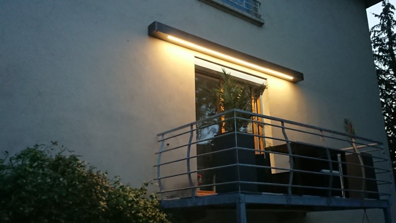 Balcon avec un luminaire lumineux, une rambarde en métal et une grande plante.