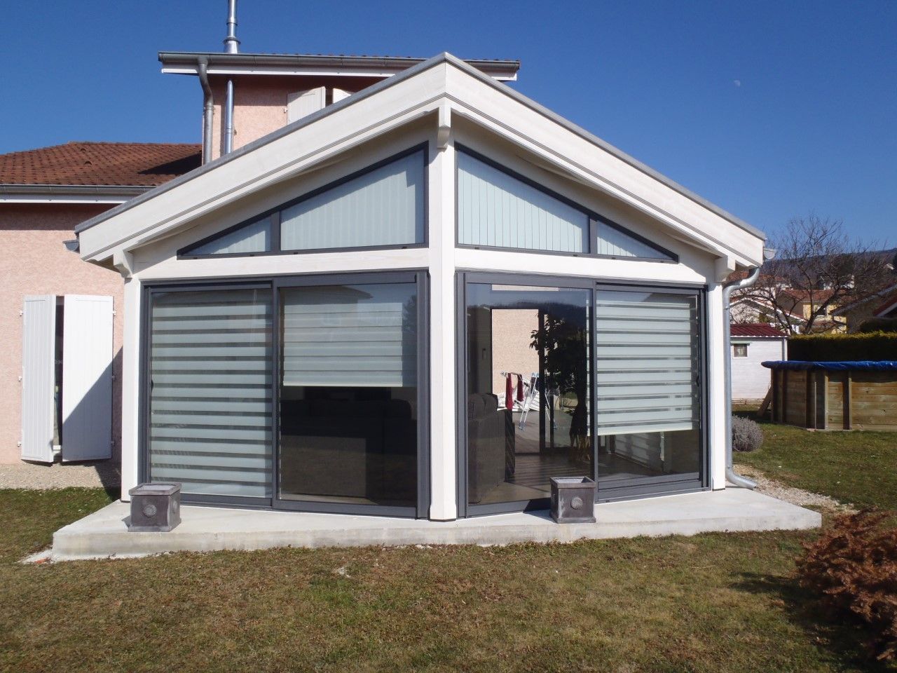 Véranda attenante à une maison au toit blanc et aux grandes fenêtres, entourée d'une pelouse ensoleillée.