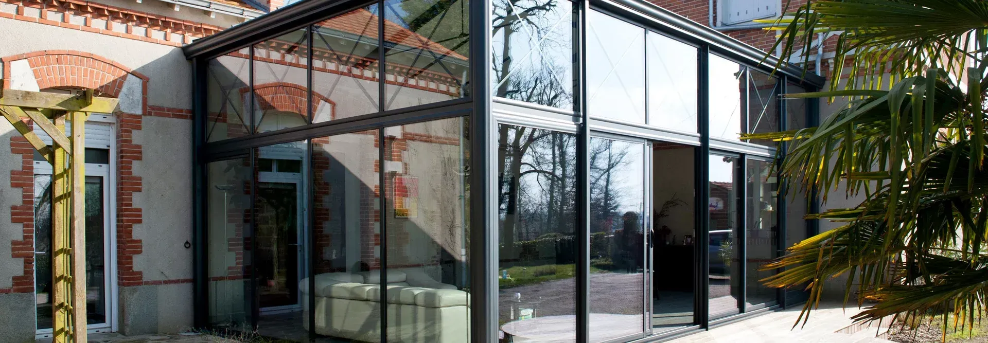Extension vitrée d'un bâtiment à façade en briques rouges, design moderne, palmier au premier plan.