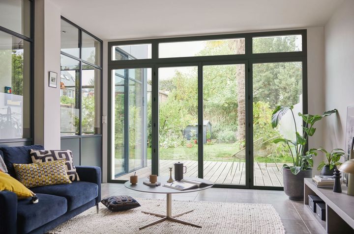 Salon avec de grandes fenêtres donnant sur un jardin. Canapé bleu, table basse, tapis et plante en pot.