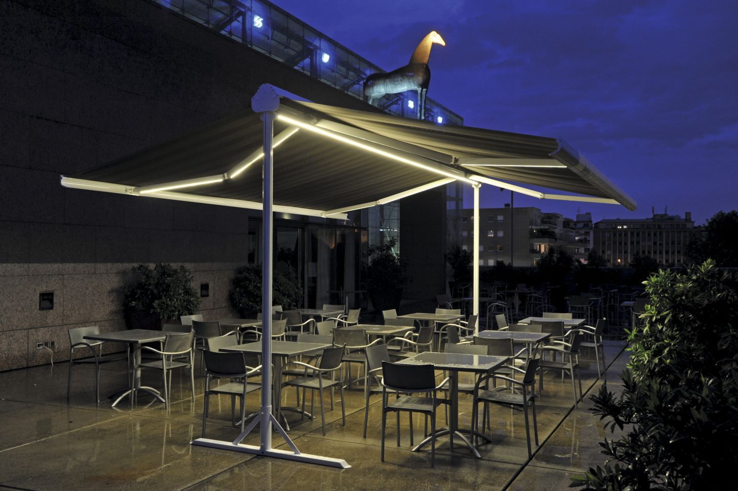 Terrasse extérieure la nuit, auvent illuminé avec tables et chaises, bâtiment illuminé en arrière-plan.