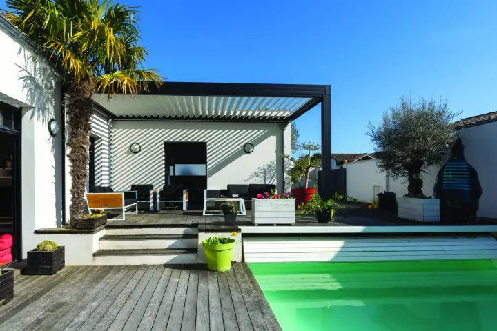 Patio au bord de la piscine avec terrasse en bois, pergola et palmier sous un ciel bleu.