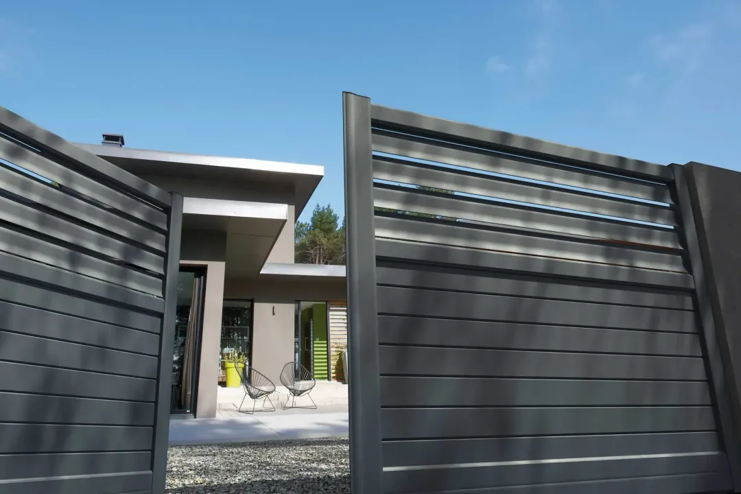 Portail à lattes horizontales grises ouvert, révélant une maison moderne avec un ciel bleu clair au-dessus.