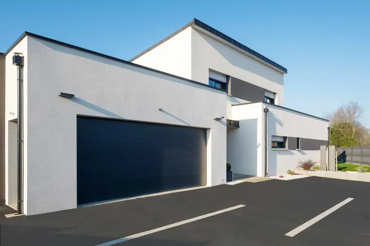 Maison blanche moderne à deux étages avec une porte de garage noire et une allée asphaltée par une journée ensoleillée.