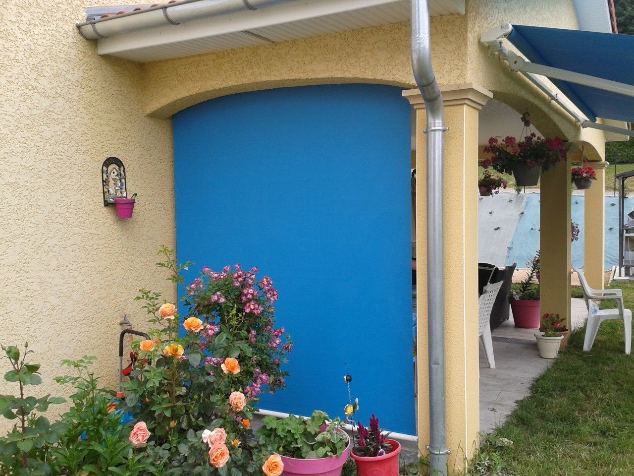 Un auvent bleu recouvre une porte d'entrée sur un bâtiment en stuc jaune. Fleurs et plantes devant.
