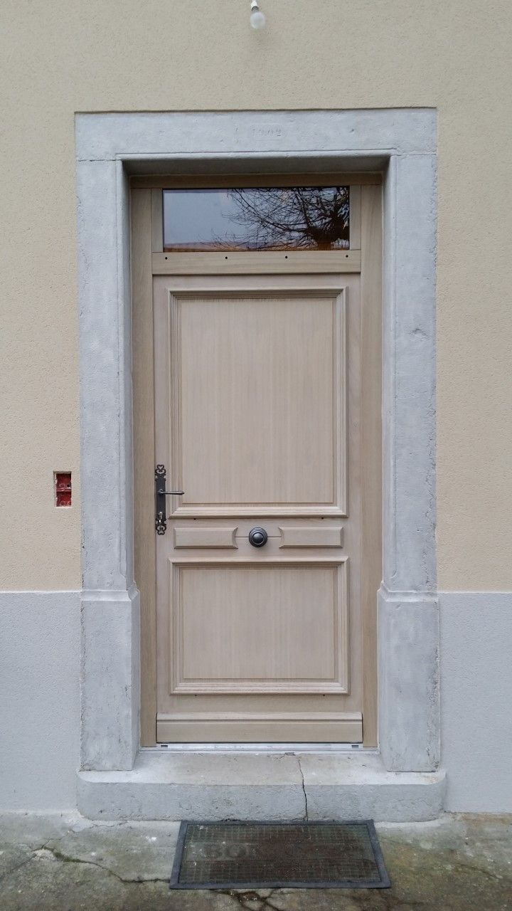 Une porte en bois clair avec une fenêtre au-dessus, encadrée de pierre, contre un mur beige.