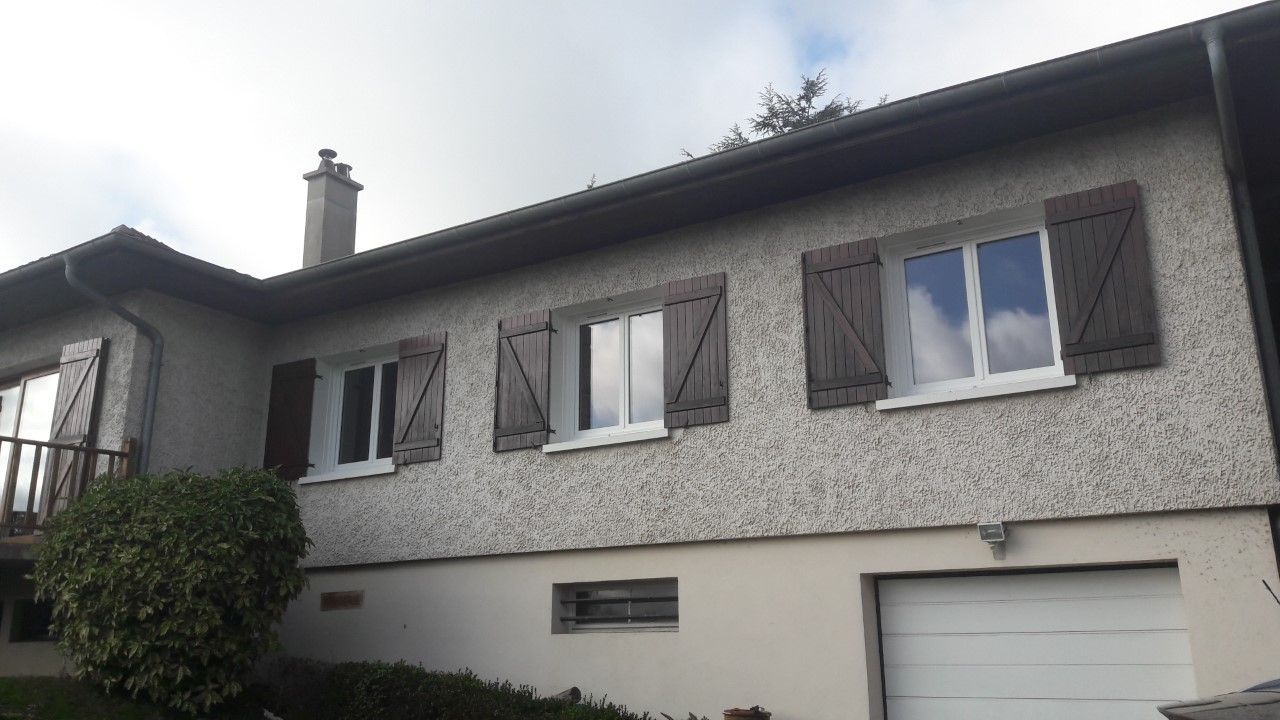 Maison beige avec des volets marron et une porte de garage blanche sous un ciel nuageux.