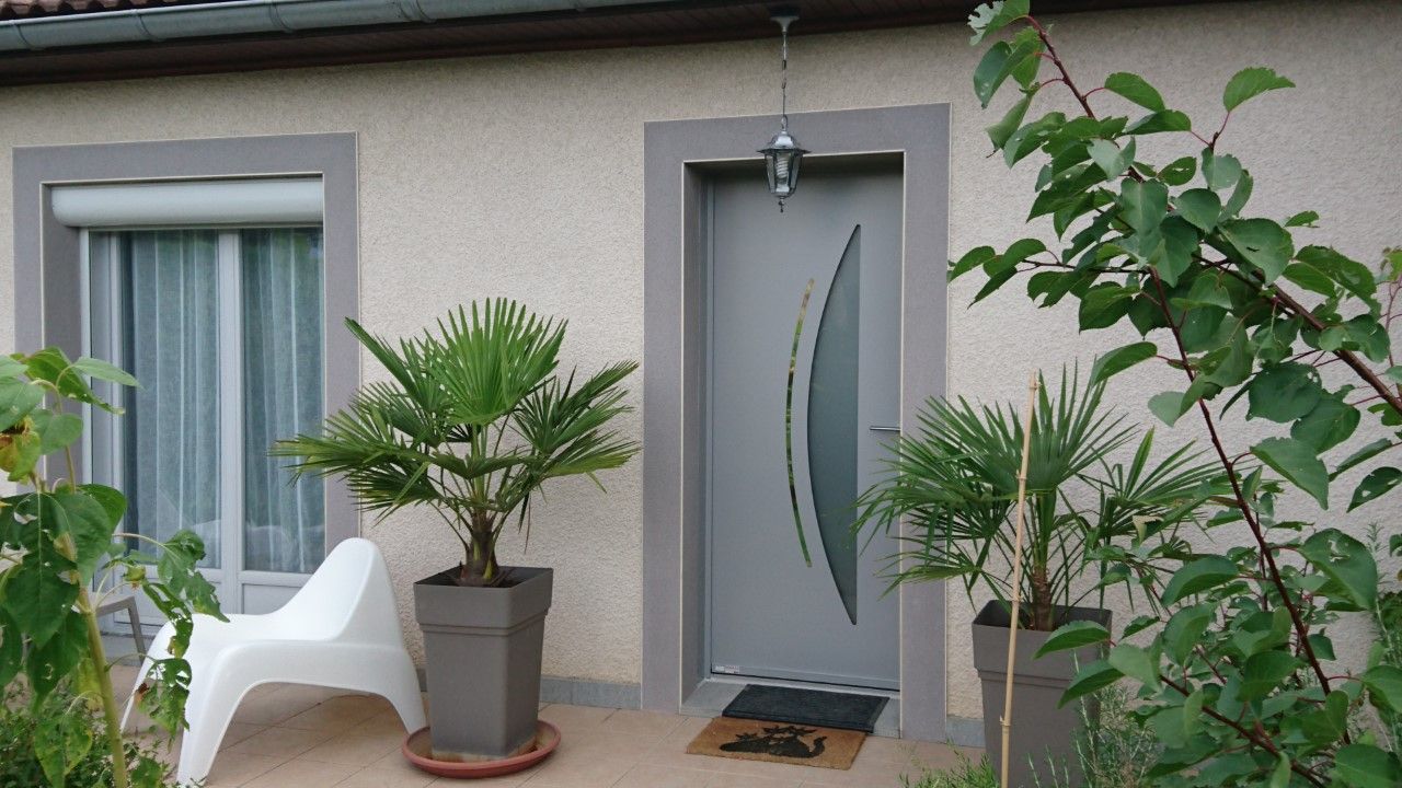 Une porte grise à vitre dépolie, flanquée de palmiers en pot, et une chaise blanche sur un petit porche.