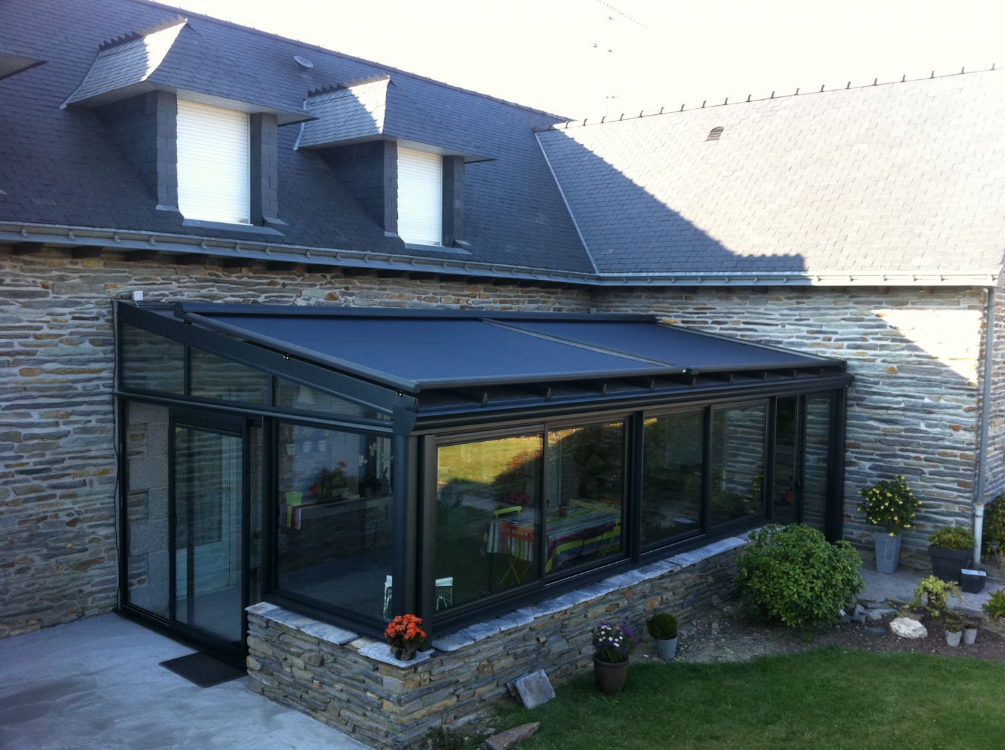Extension de type véranda avec toit noir et parois vitrées, attenante à une maison aux murs en pierre.