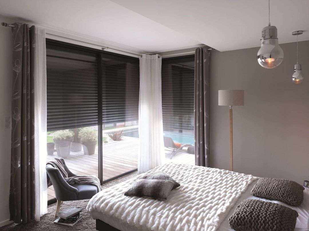 Chambre avec de grandes fenêtres, des stores foncés, des rideaux blancs transparents, des rideaux à motifs gris et un lit.