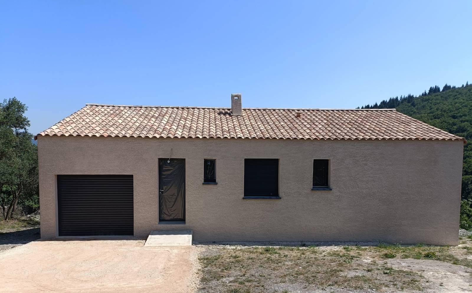 Une maison de plain-pied couleur sable, avec une porte de garage foncée, une porte d'entrée et trois fenêtres sous un toit de tuiles, se détachant sur un ciel dégagé.