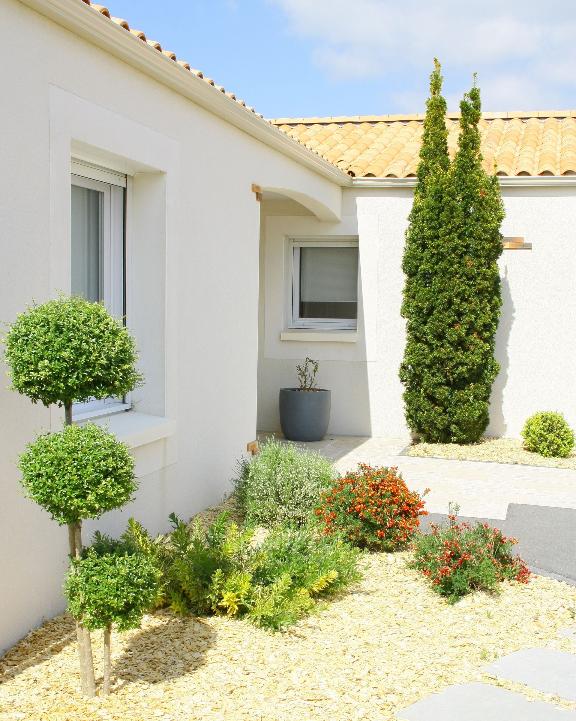 Un coin de maison aux tons clairs, avec une cour gravillonnée agrémentée d'un arbre topiaire, de divers arbustes et d'un grand conifère colonnaire.