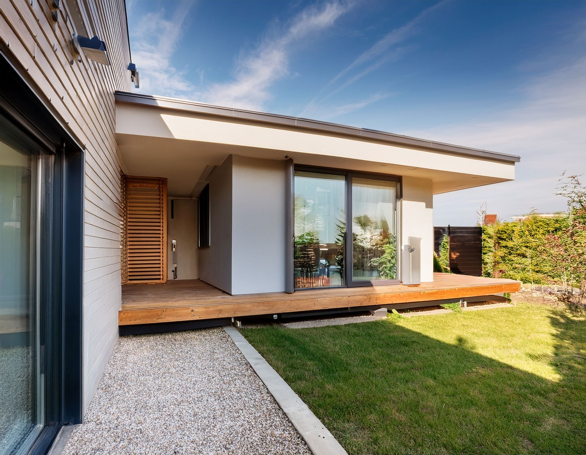 Une maison moderne avec une terrasse en bois, un chemin de gravier et une pelouse bien entretenue sous un ciel bleu azur.