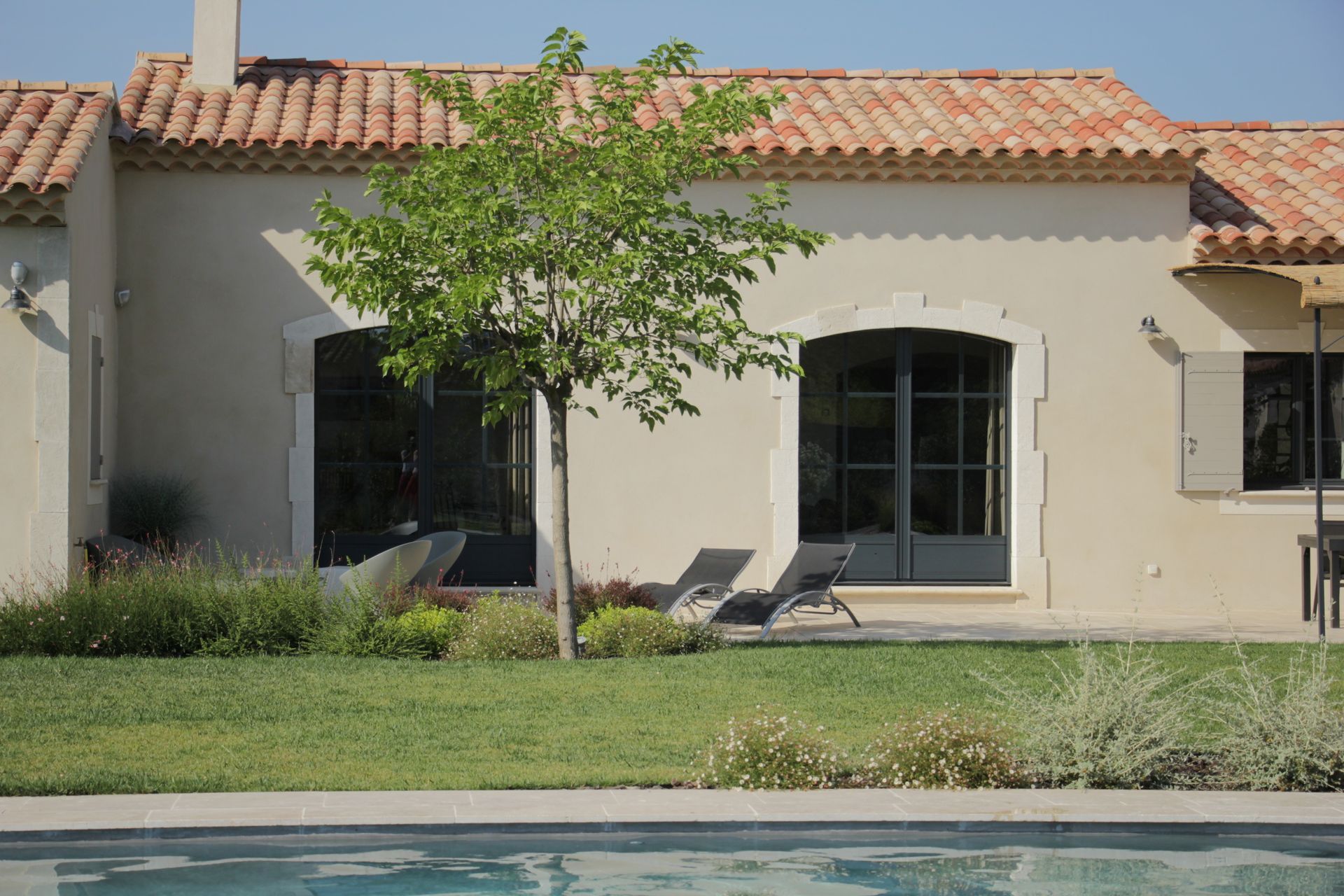 Une maison beige au toit de tuiles se dresse derrière une pelouse verdoyante, un arbre et une piscine dans le jardin.