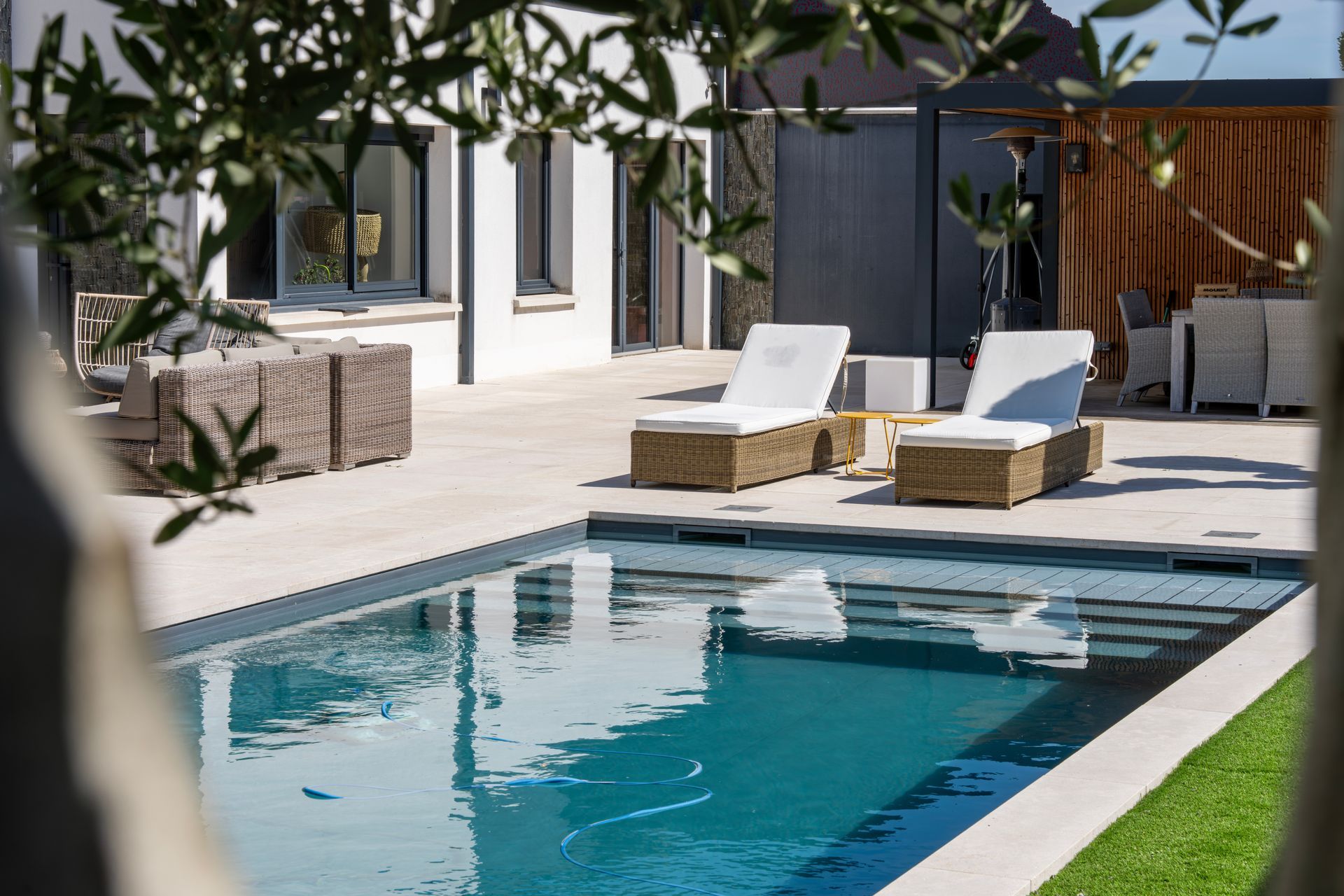 Une piscine avec deux chaises longues sur une terrasse en pierre, à côté d'une maison moderne et d'un espace repas extérieur.