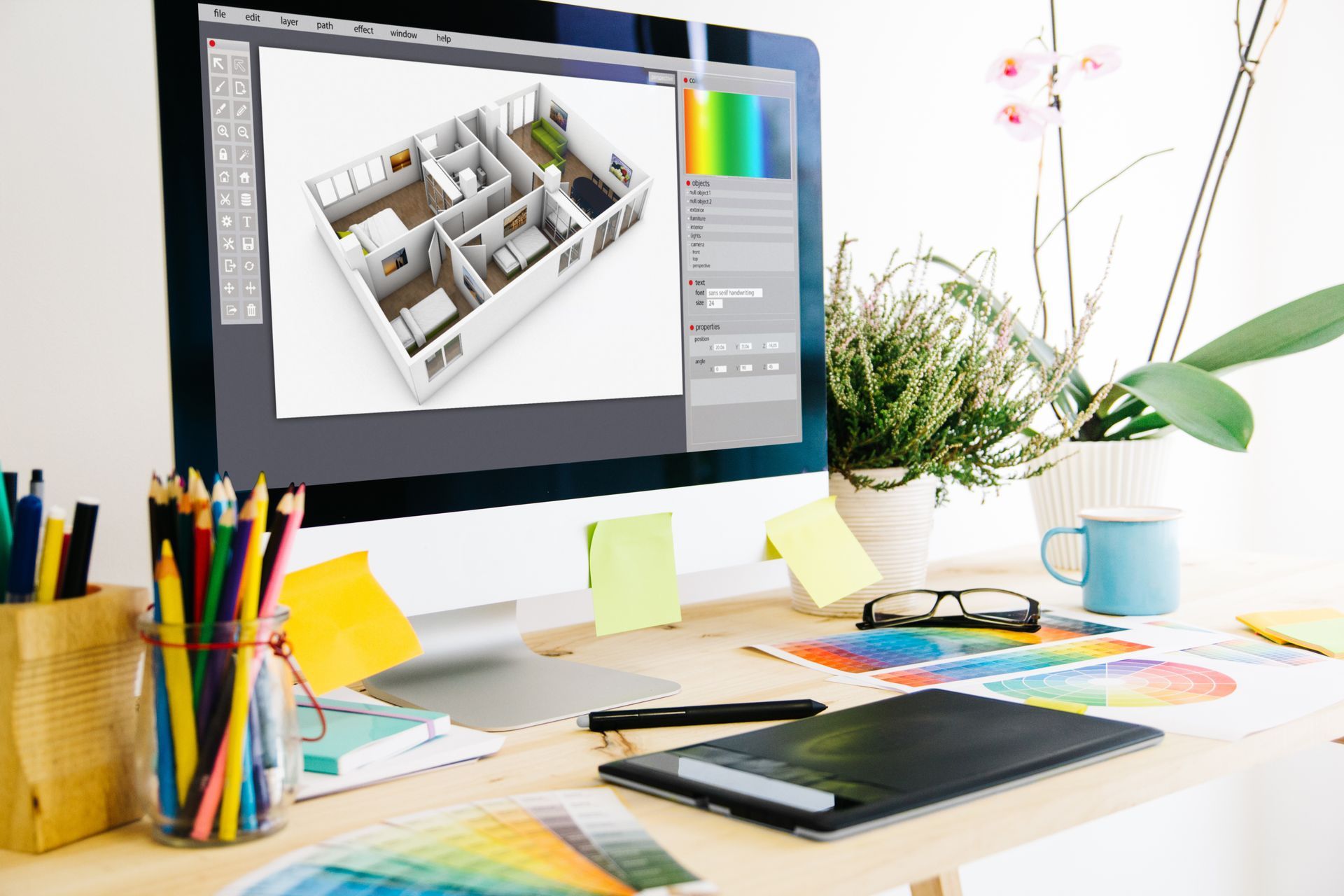 Un écran affiche un plan d'étage en 3D posé sur un bureau avec des crayons de couleur, une tablette graphique, une plante et des échantillons de couleurs.