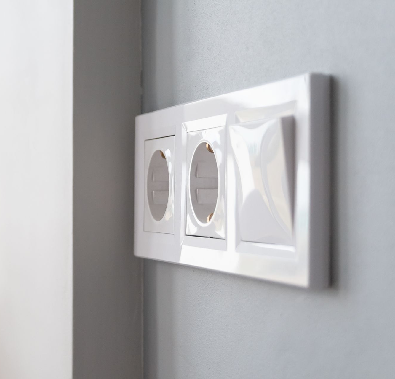 Vue latérale d'une plaque murale blanche horizontale contenant deux prises de courant et un interrupteur simple, sur un mur gris.
