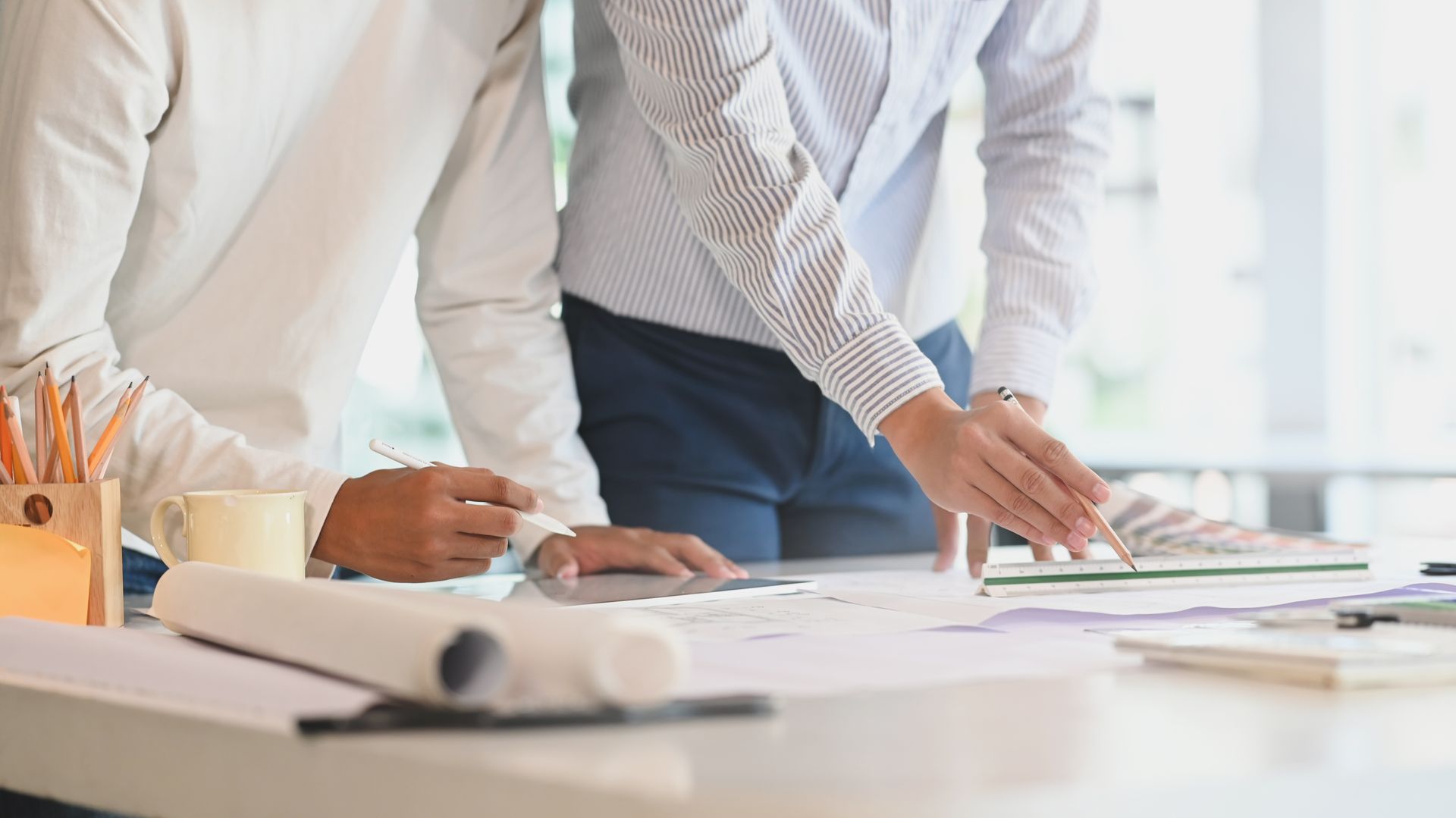 Deux professionnels examinent et désignent du doigt des plans architecturaux posés sur un bureau lumineux rempli d'outils de dessin.