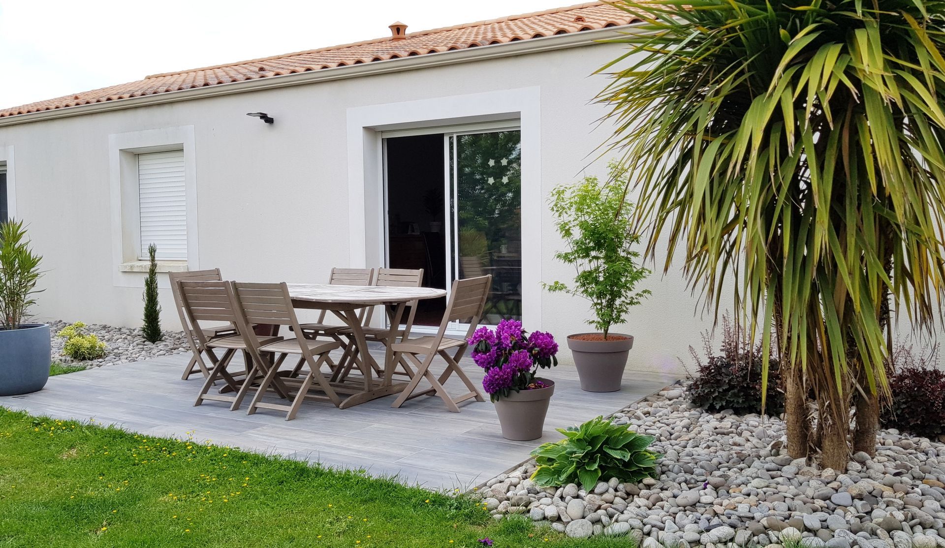 Une terrasse avec une table et des chaises en bois devant une maison blanche avec une pelouse et des plantes en pot.