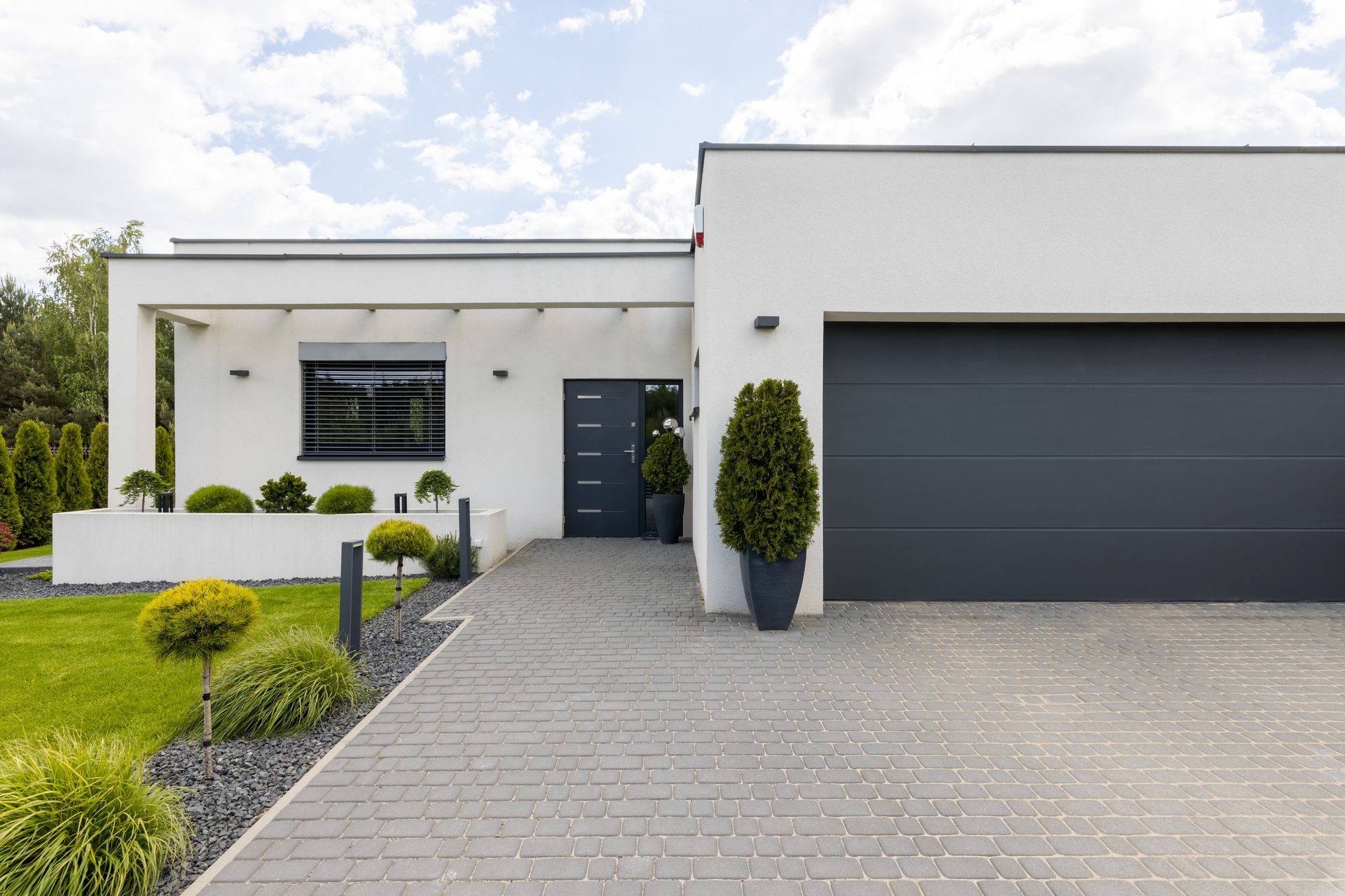Une maison moderne de plain-pied, blanche, avec une porte de garage grise, une allée pavée et un aménagement paysager verdoyant et soigné.