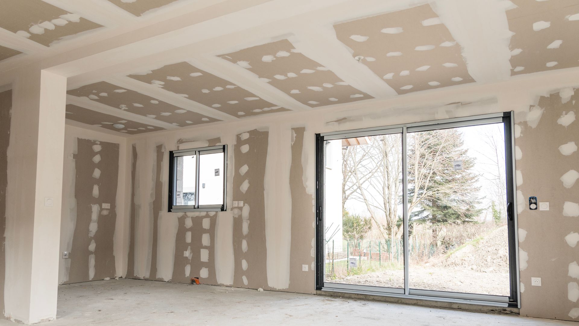 Intérieur d'une pièce en construction avec des cloisons sèches non finies, des joints apparents, une fenêtre et une porte coulissante en verre.