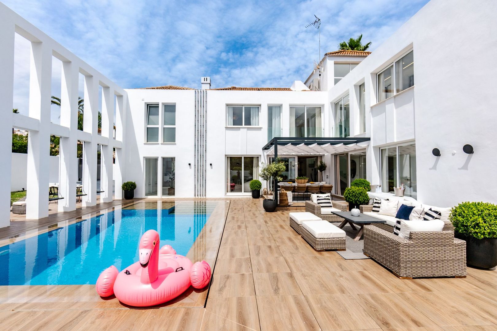 Une terrasse extérieure ensoleillée avec une piscine rectangulaire, un flamant rose gonflable et une architecture de villa blanche moderne.