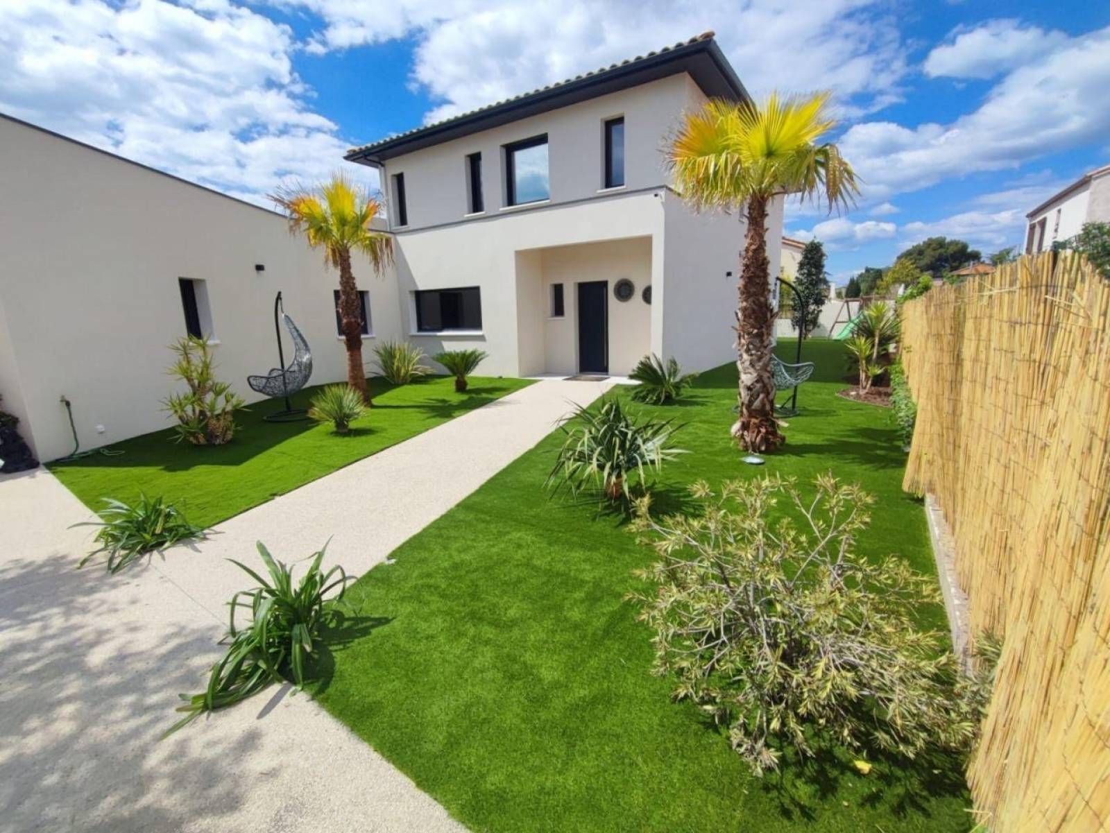 Une maison moderne à deux étages avec une façade beige, agrémentée d'un chemin de pierre menant à l'entrée à travers un jardin paysager.