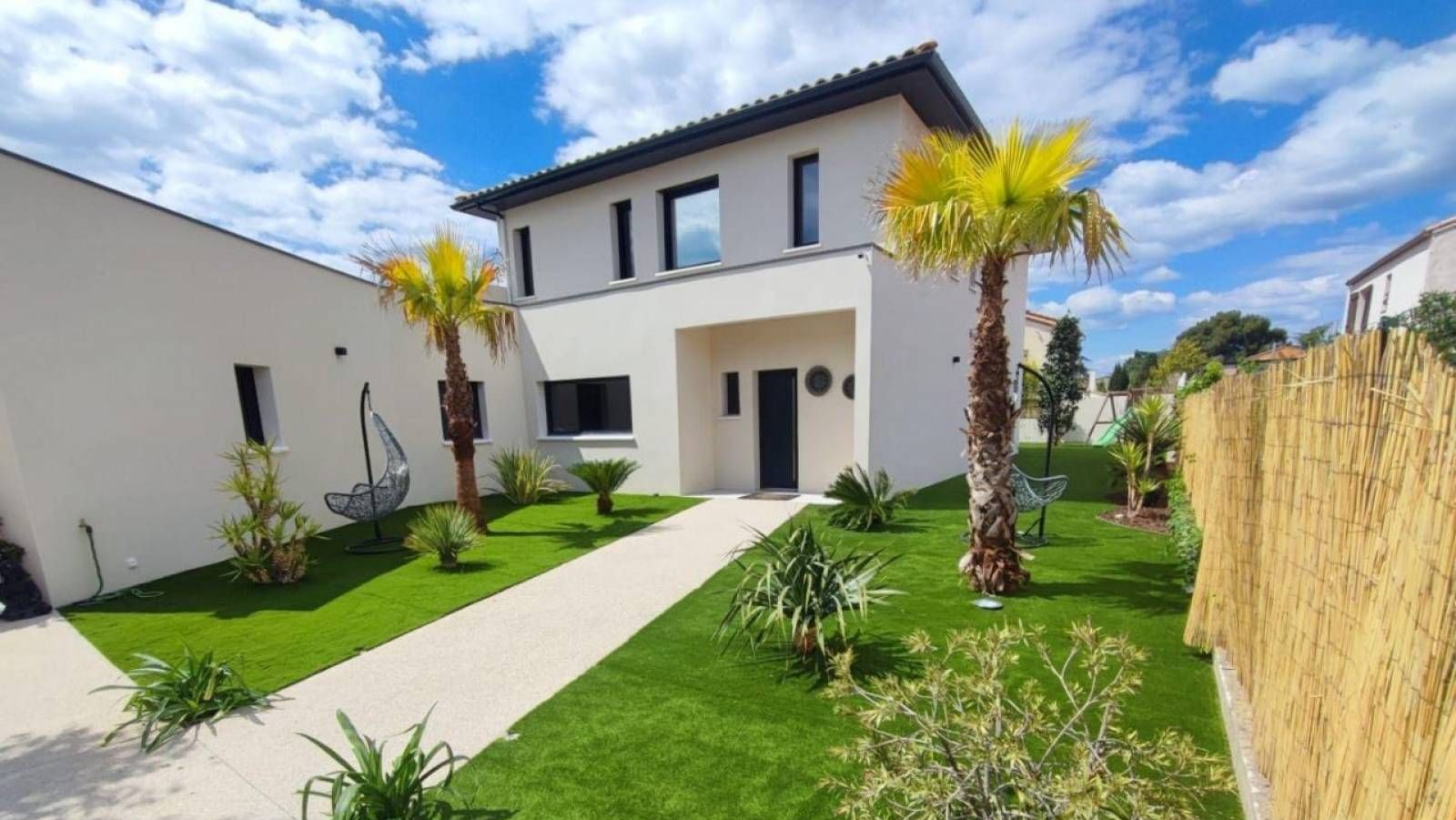 Une maison moderne de deux étages aux tons clairs, avec un chemin traversant une pelouse verte, deux palmiers et une clôture en bambou sur le côté.
