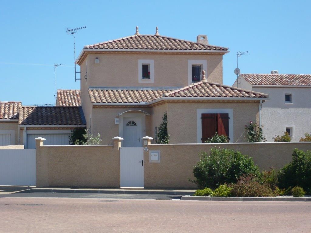 Une maison beige à deux étages avec un toit en tuiles de terre cuite, des boiseries blanches et un muret, vue depuis une rue résidentielle.