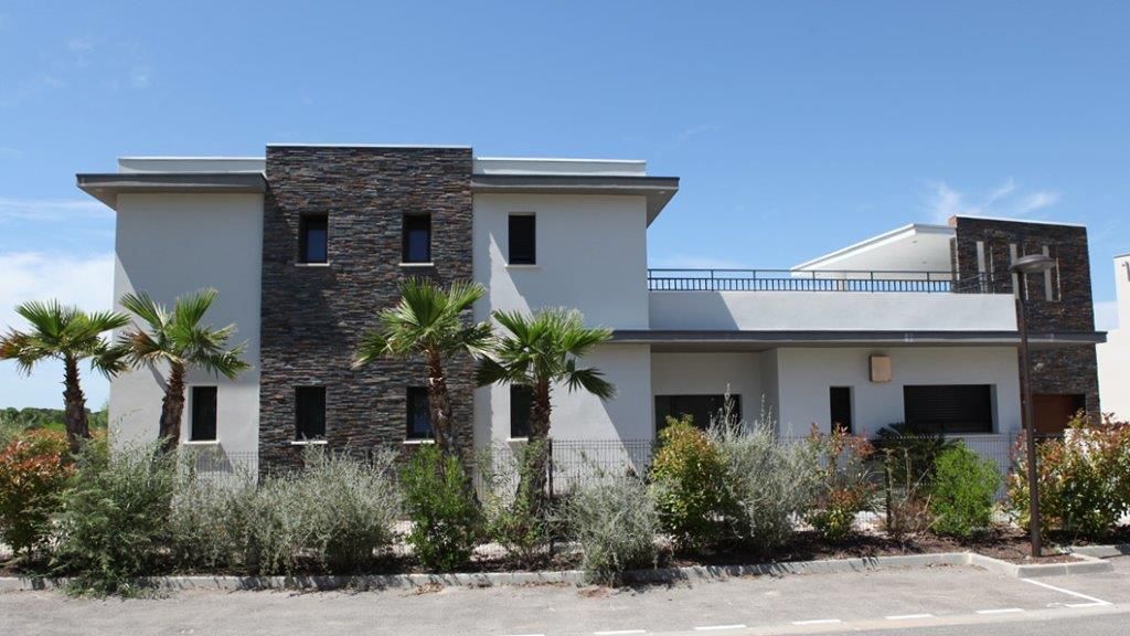 Une maison moderne à deux étages avec des murs gris clair, des touches de pierre foncée et un jardin de devant avec des palmiers sous un ciel bleu.