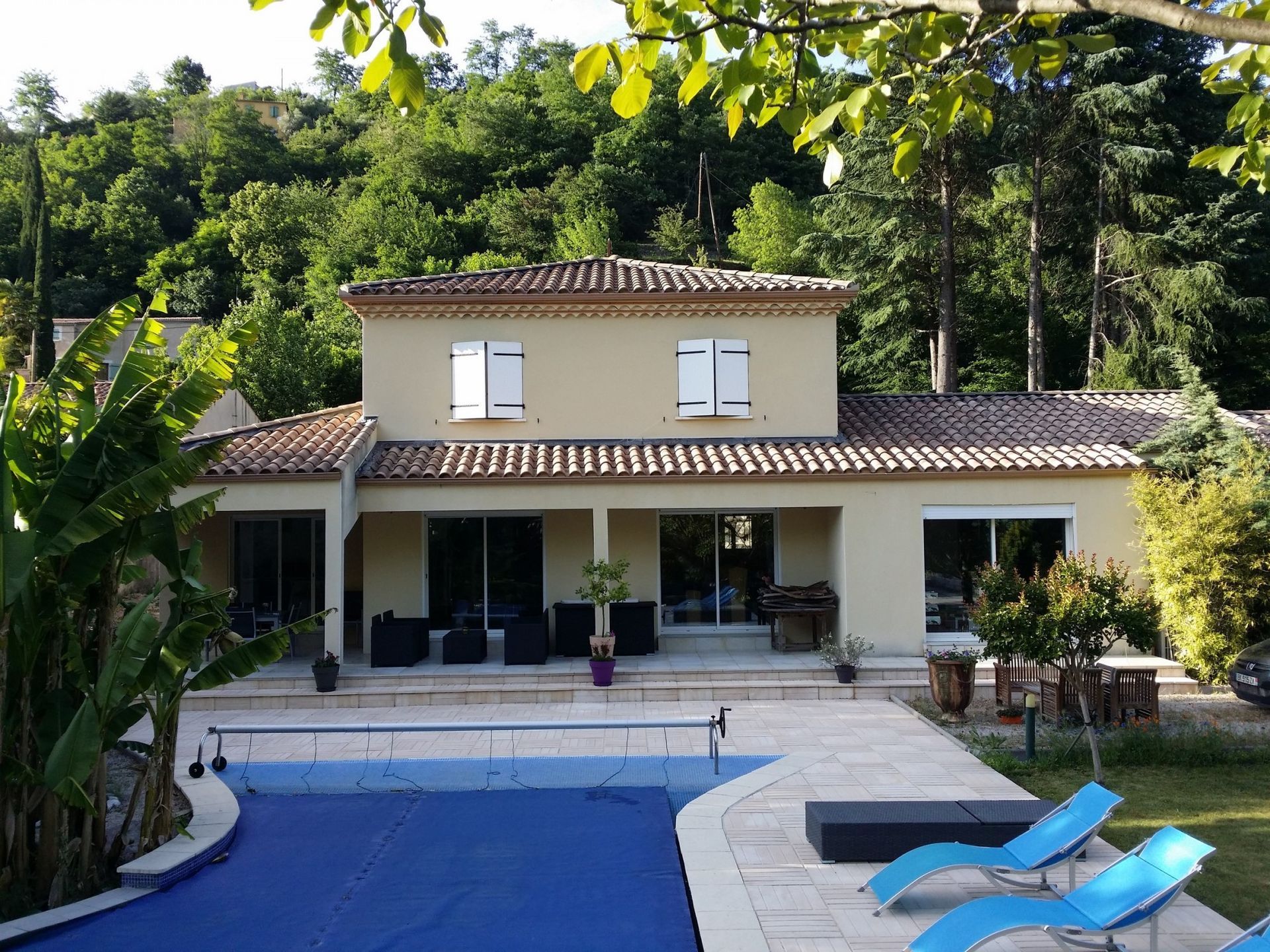 Par une journée ensoleillée, une maison beige à deux étages avec des volets blancs se dresse derrière une piscine recouverte d'une bâche bleue.