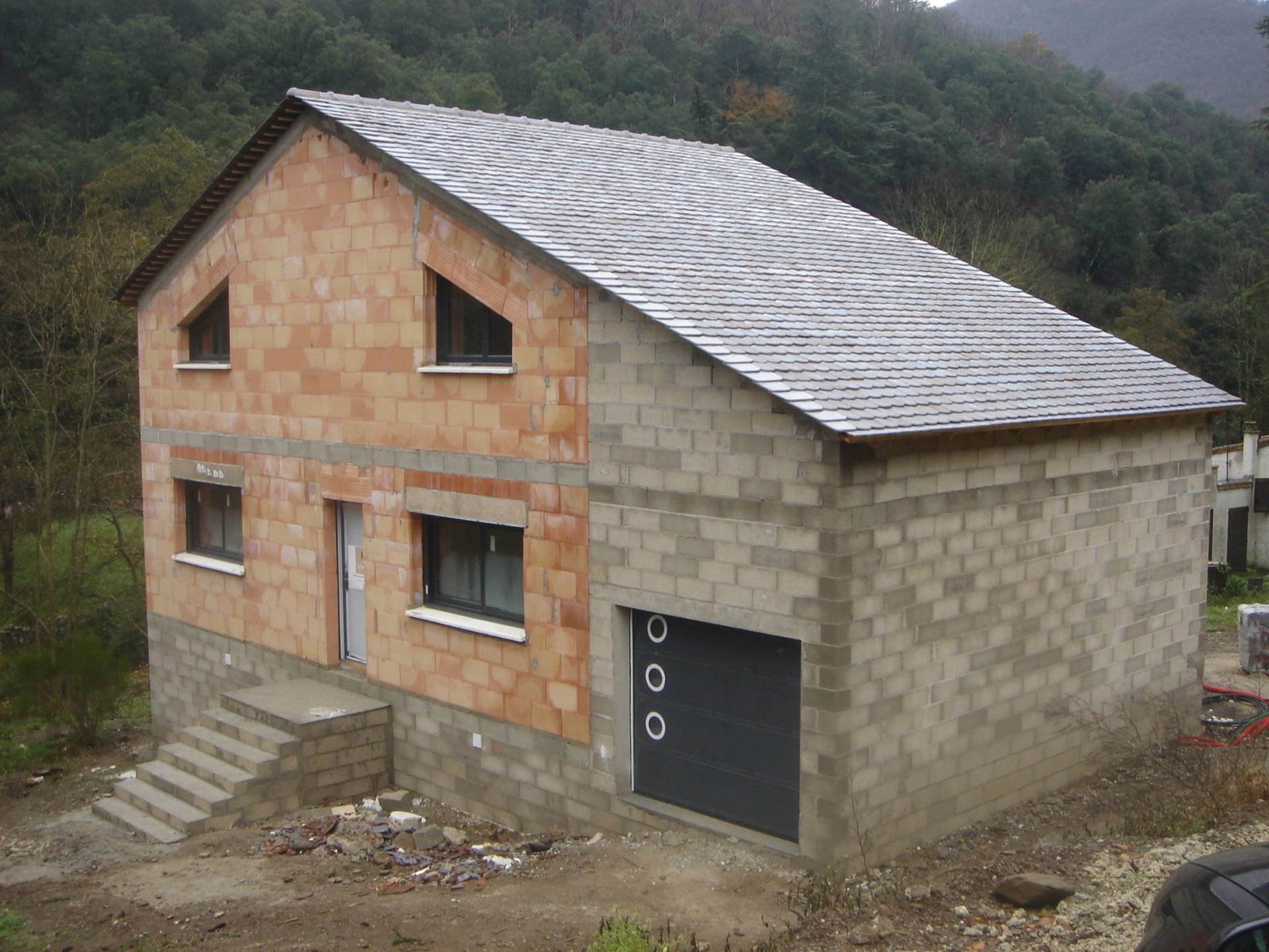 Maison à deux étages en construction, avec des murs en briques et des blocs de béton gris, un toit sombre et un garage, située en milieu rural.