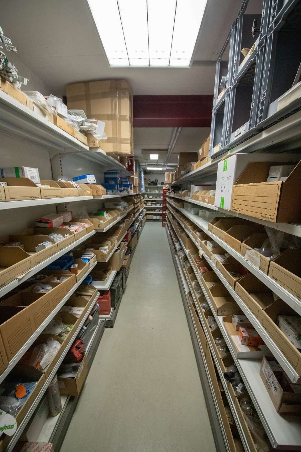 Schmaler Lagergang mit Regalen voller Kisten und Teile.