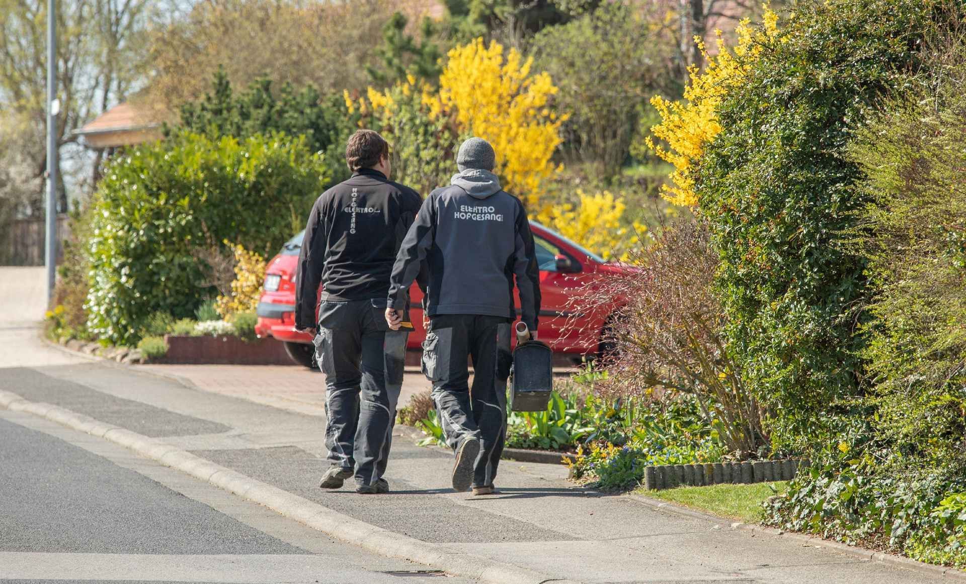 Zwei Personen gehen auf einem Bürgersteig an einem roten Auto und Büschen vorbei. Einer trägt eine blaue Jacke, der andere eine schwarze Jacke.