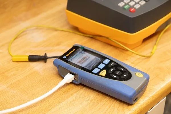 Blau-graues Netzwerktestgerät mit Bildschirm, Ethernet-Kabel und gelbem Kabel, verbunden mit einem schwarz-gelben Testgerät auf einer Holzoberfläche.