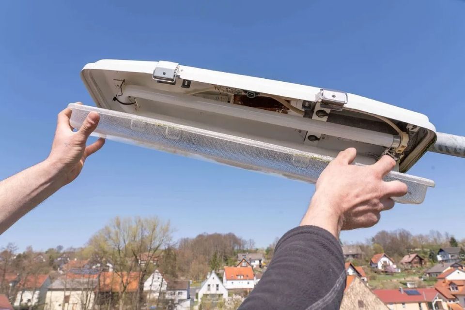 Hände halten vor blauem Himmel eine durchsichtige Plastikabdeckung einer Straßenlaterne offen.