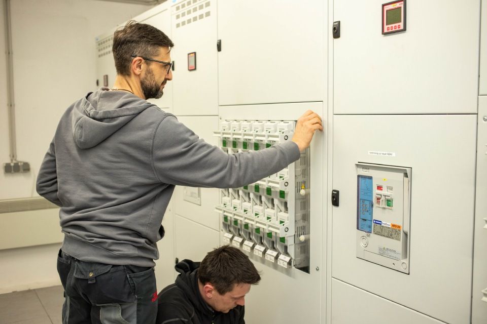 Zwei Personen arbeiten an Schalttafeln in einem Hauswirtschaftsraum. Einer steht und stellt einen Schalter ein, der andere kniet.