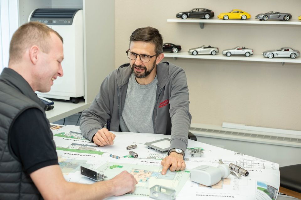 Zwei Männer sitzen mit Bauplänen an einem Tisch und besprechen ein Projekt. Im Hintergrund stehen Miniaturautomodelle auf einem Regal.